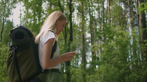 A-young-woman-with-a-mobile-phone-walks-through-the-forest-traveling-with-a-backpack-in-slow-motion.-Traveler-in-shorts-in-the-woods-looking-for-gps-satellites